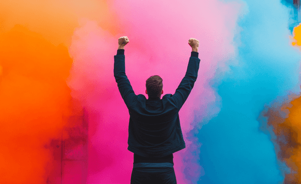 Person with raised arms stands before vibrant orange, pink, and blue smoke clouds, creating a dynamic and colorful scene.