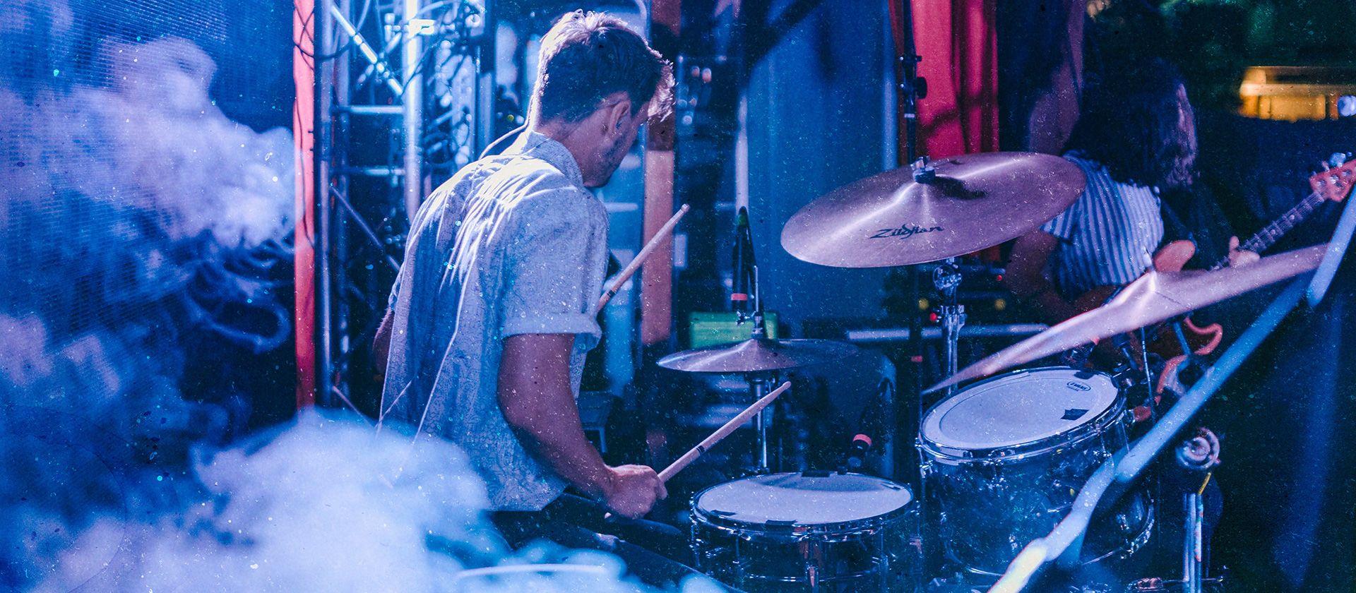 Man playing the drums in blue light amongst smoke on stage.