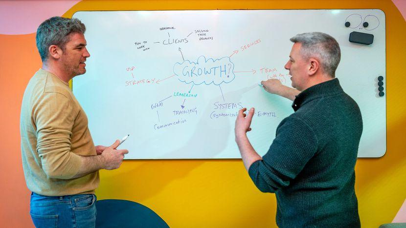 Two men conversing in front of a whiteboard.